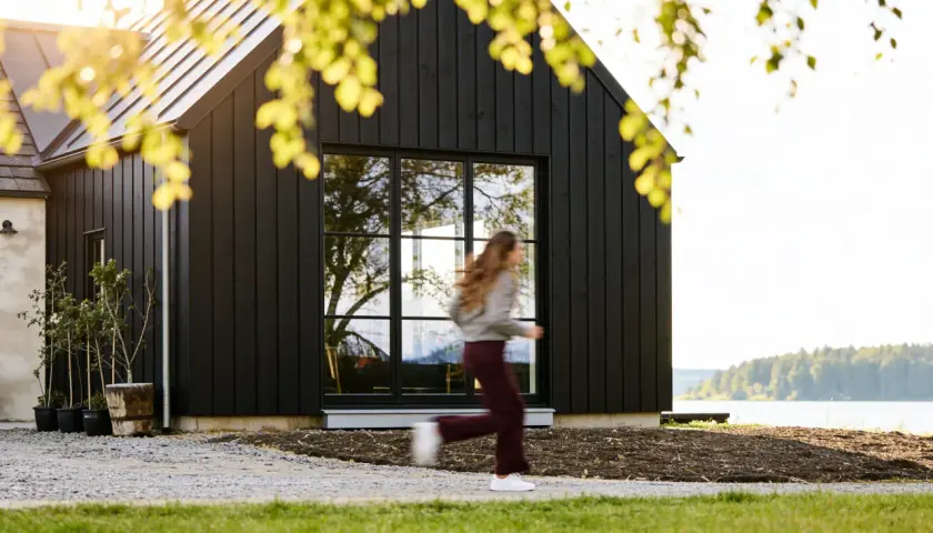 Svart trähus med stora fönster vid en sjö. En person springer förbi. Utbyggnad hus inspiration för moderna hem.