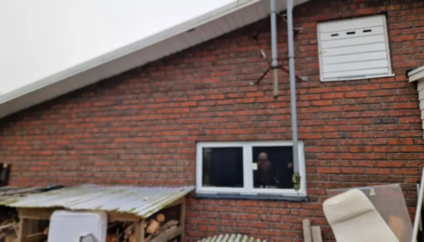 Röd tegelfasad konstruktion med ett fönster och en vit garageport. Vedhögar och möbler syns framför huset.