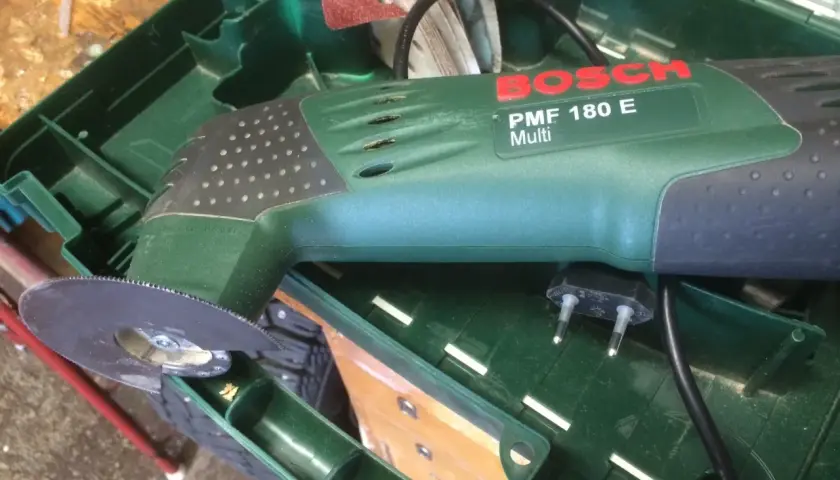 Bosch PMF 180 E multiverktyg med sågblad redo att kapa plasttak. Tillbehör och laddare ligger i lådan.
