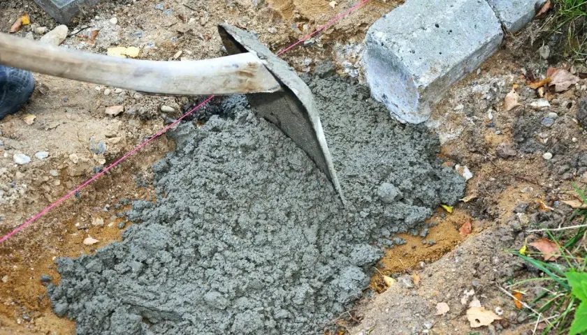 En hacka sprider jordfuktig betong i en grop. Betongblock och en rosa snodd syns i bakgrunden.