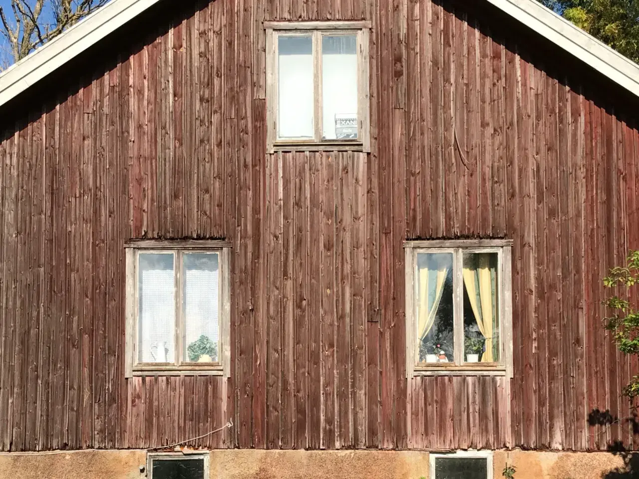 Rödbrun panel på ett gammalt timmerhus med tre fönster. Ett fönster högst upp har en skylt.