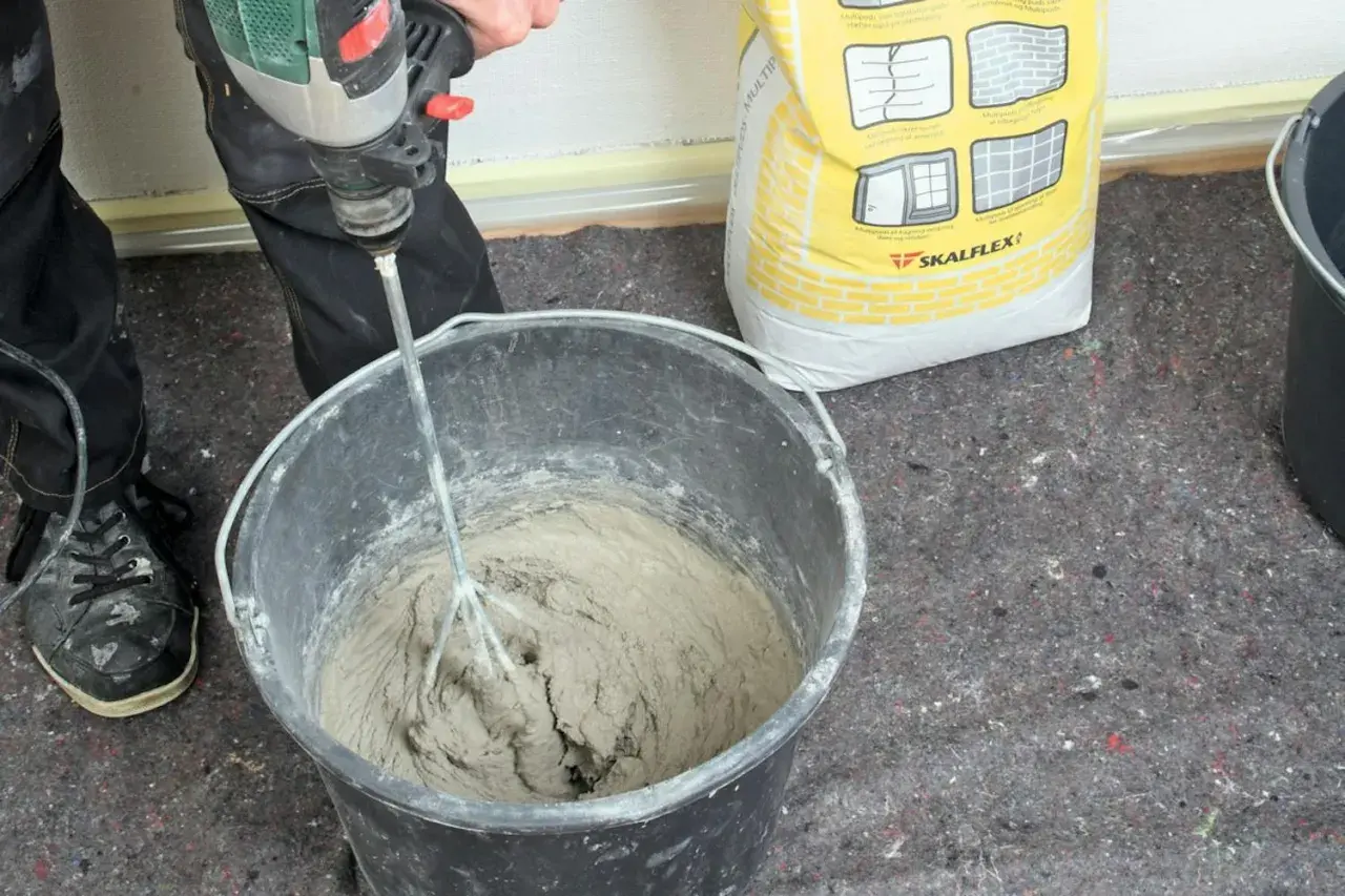 En person använder en borrmaskin för att blanda betong i en hink. En gul säck med Skalflex-märkning står bredvid.