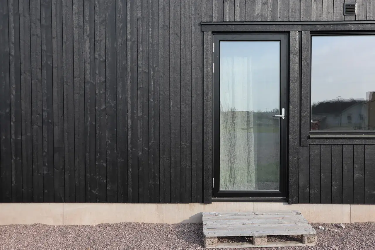 Svart husgrund med träpanel, glasdörr och fönster. En pall som trappa leder upp.