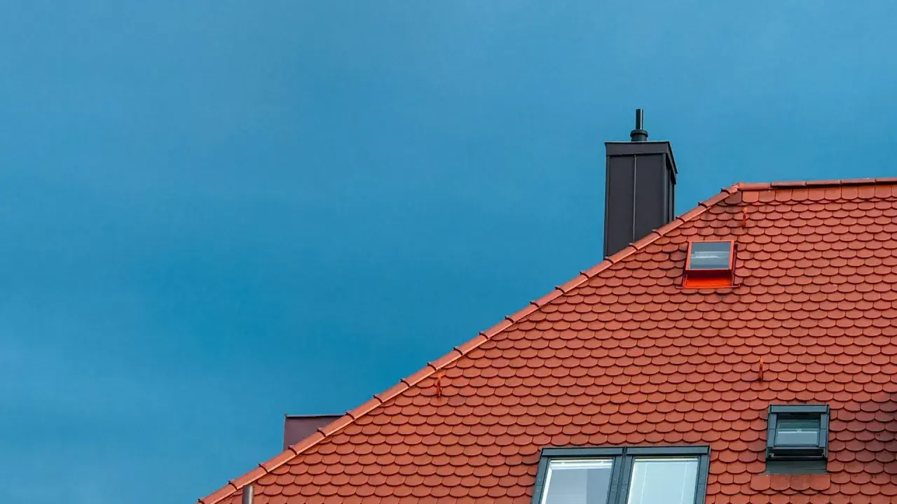 Rött tegeltak med skorsten och takfönster mot en blå himmel. Dags att byta tak?