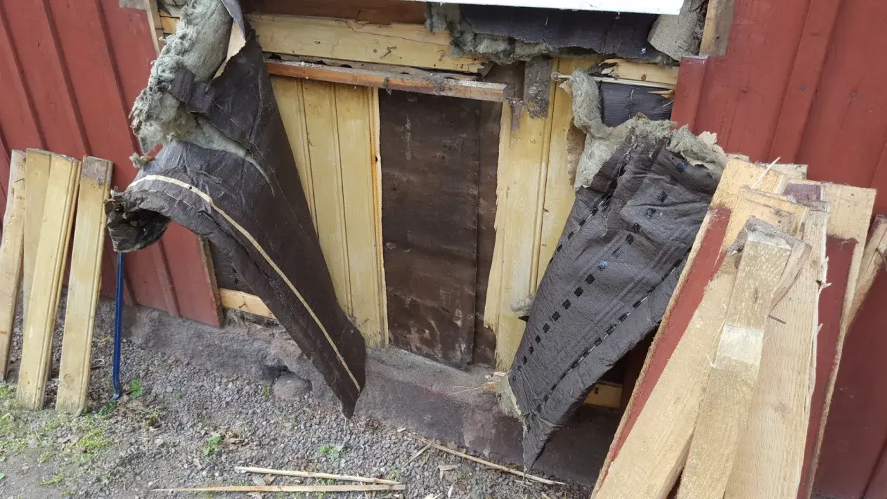 Rivning av gammalt tak. Synlig isolering och trasig vindpapp. Bräder och spillved ligger staplade.