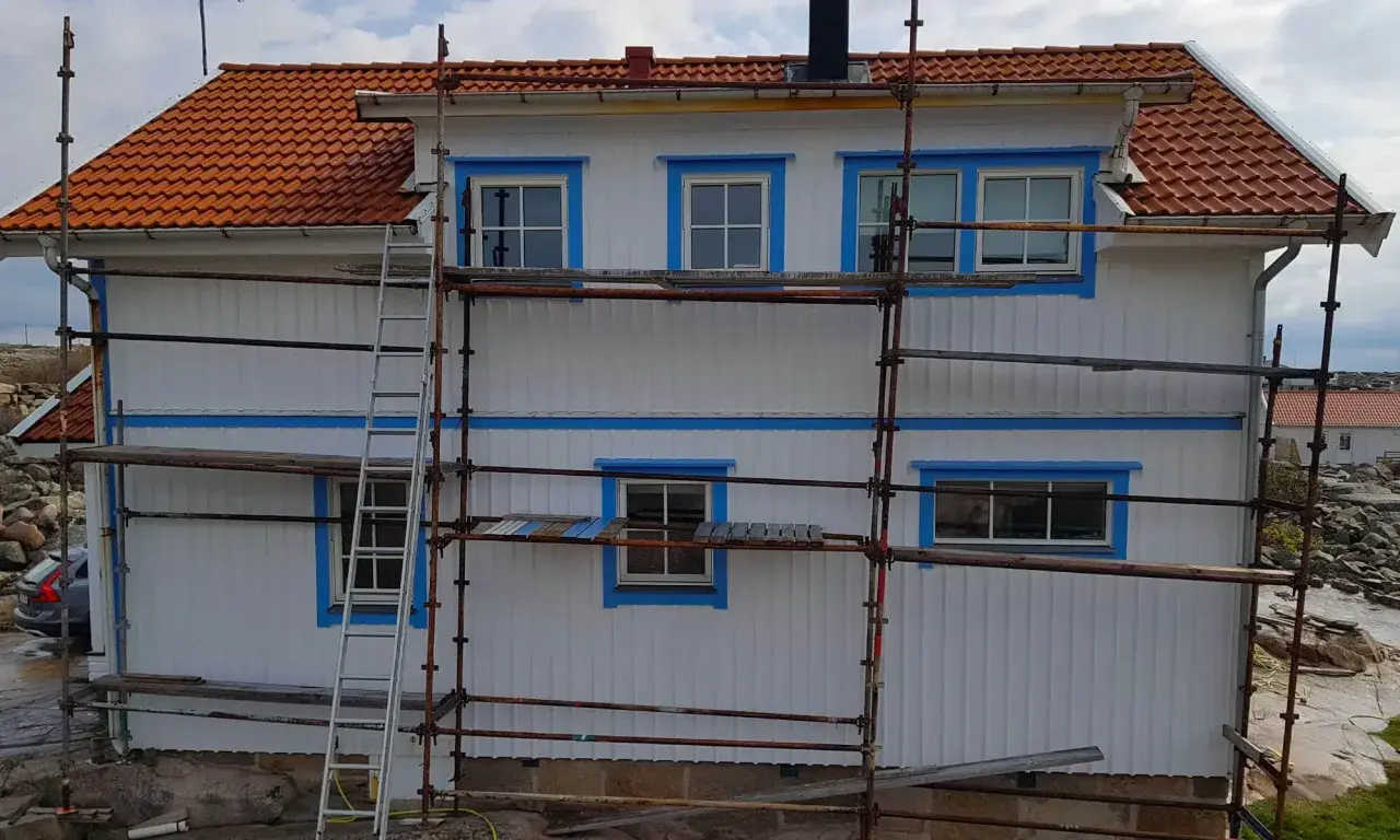 Ställningar runt ett vitt hus med orange tak. Arbetet med att isolera gammalt hus utifrån pågår.