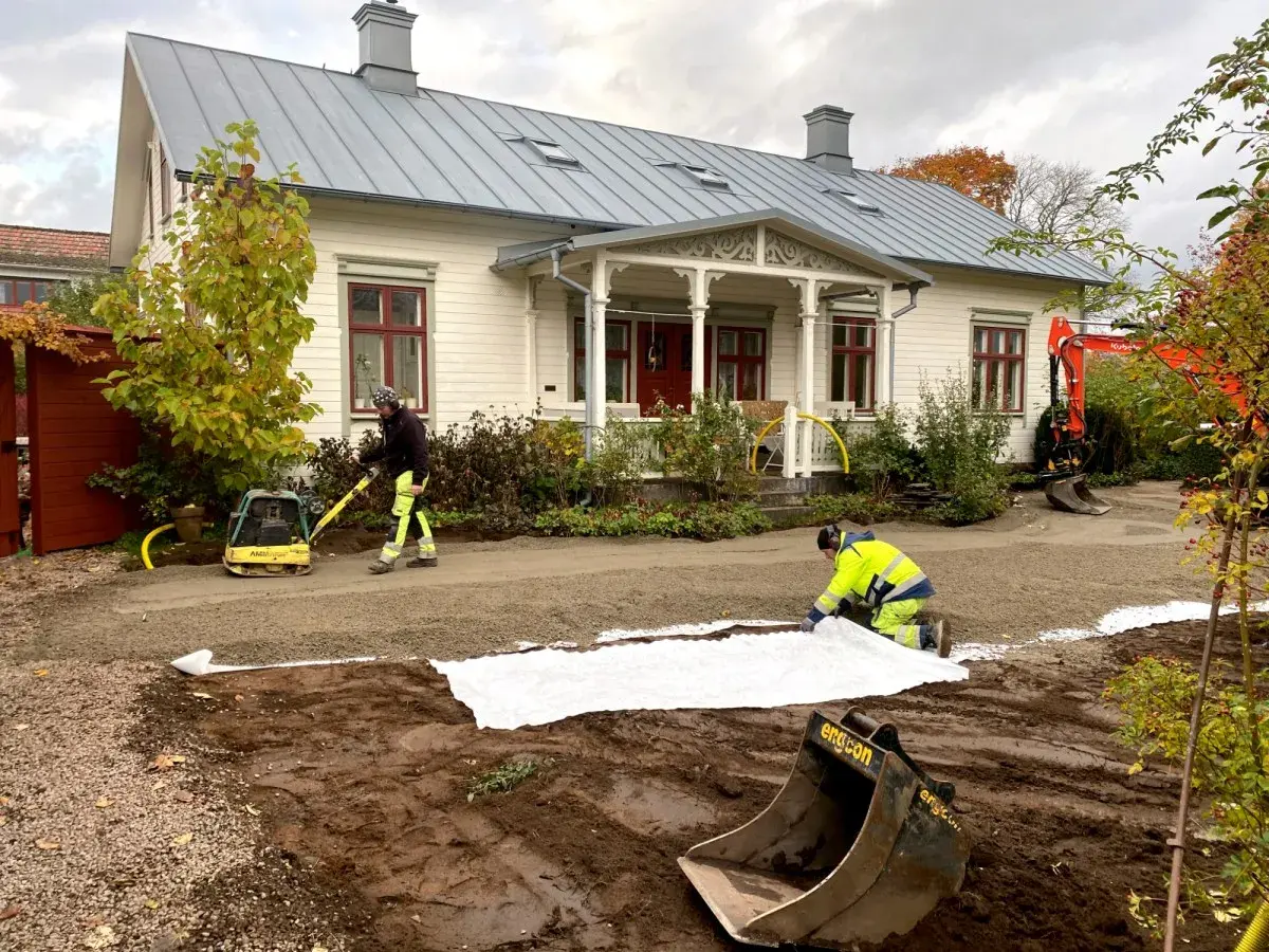 Arbetare lägger ut duk och packar grus för en gammaldags grusgång framför ett vitt hus. En grävmaskin syns i bakgrunden.