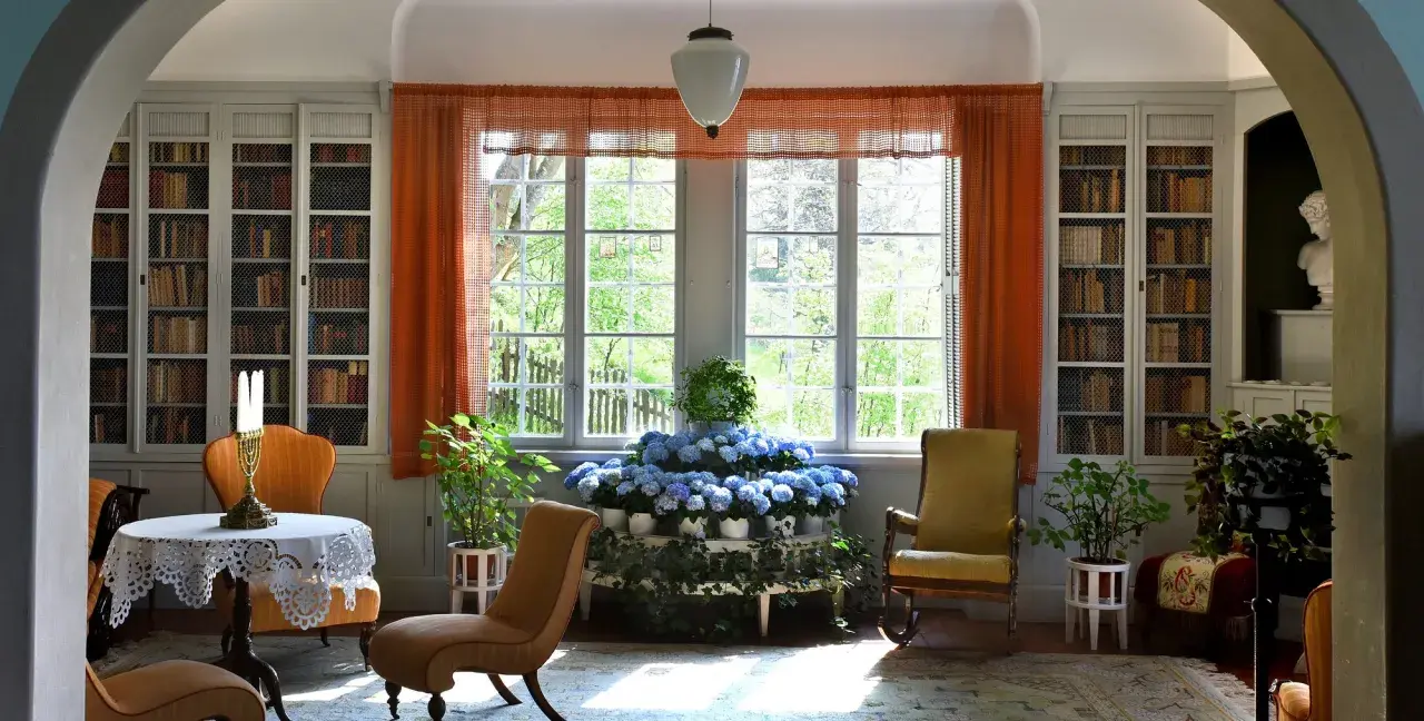 Ett rum med bokhyllor, en rund dukförsedd bord, stolar och en blomsteruppsättning. Ellen Keys inredning präglas av ljus och natur.