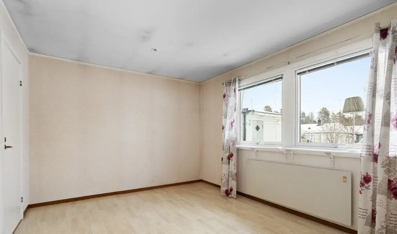 Ett rum med ljusa väggar och trägolv. Svart sot i taket syns ovanför dörren och längs kanterna. Fönster med blommig gardin och utsikt över snöklädda hus.