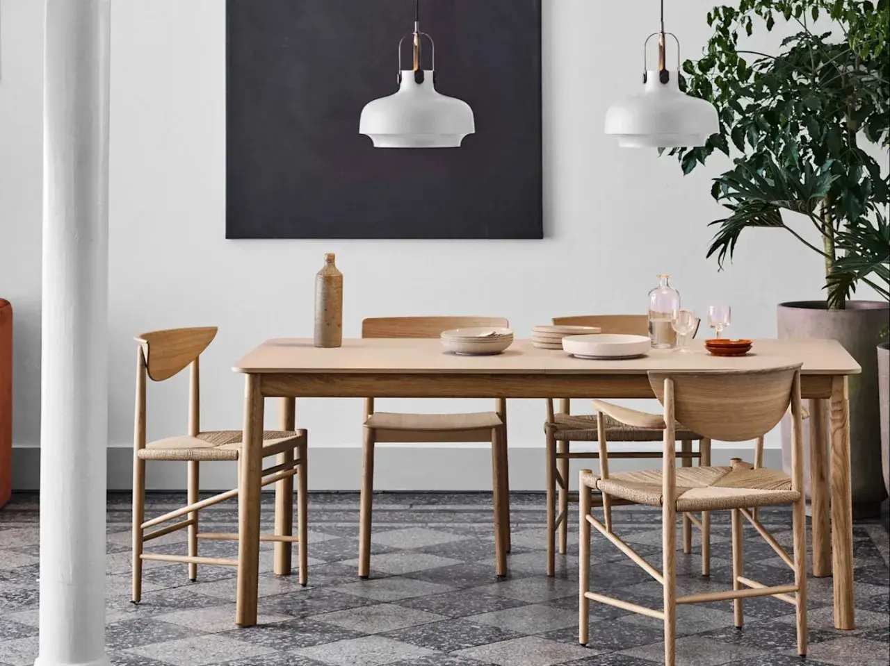 Ett ljust träbord med stolar i naturmaterial, en vas och tallrikar. Lamporna ovanför och den stora gröna växten bidrar till en känsla av antroposofisk inredning.