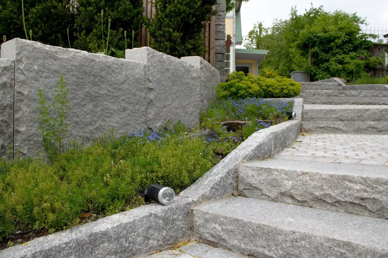 En imponerande mur av granit sten ramar in en lummig rabatt med blå blommor. En trappa i samma granit sten leder uppåt.