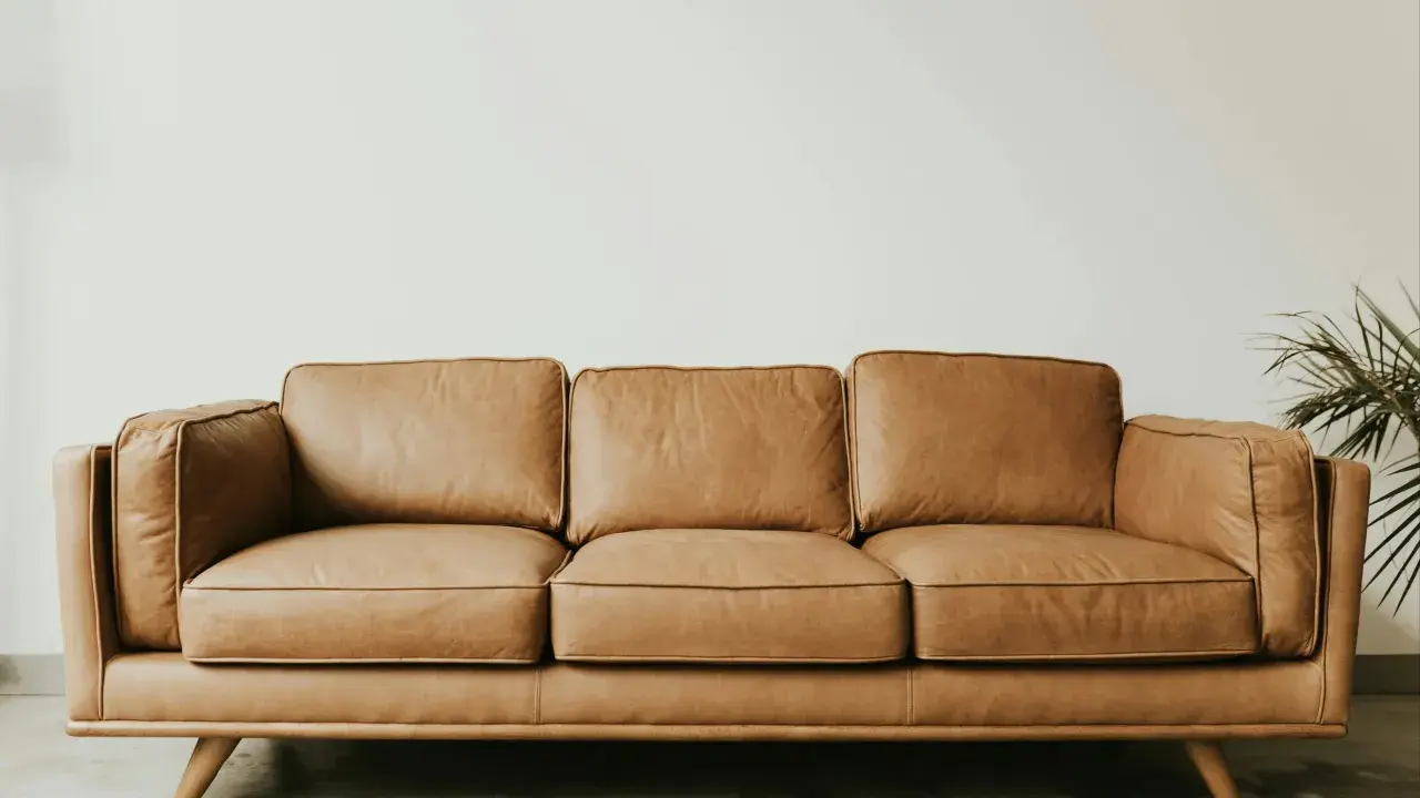 Brun lädersoffa med tre sittkuddar och ryggkuddar, på sneda träben. En grön växt syns till höger. Perfekt för hållbara hus.