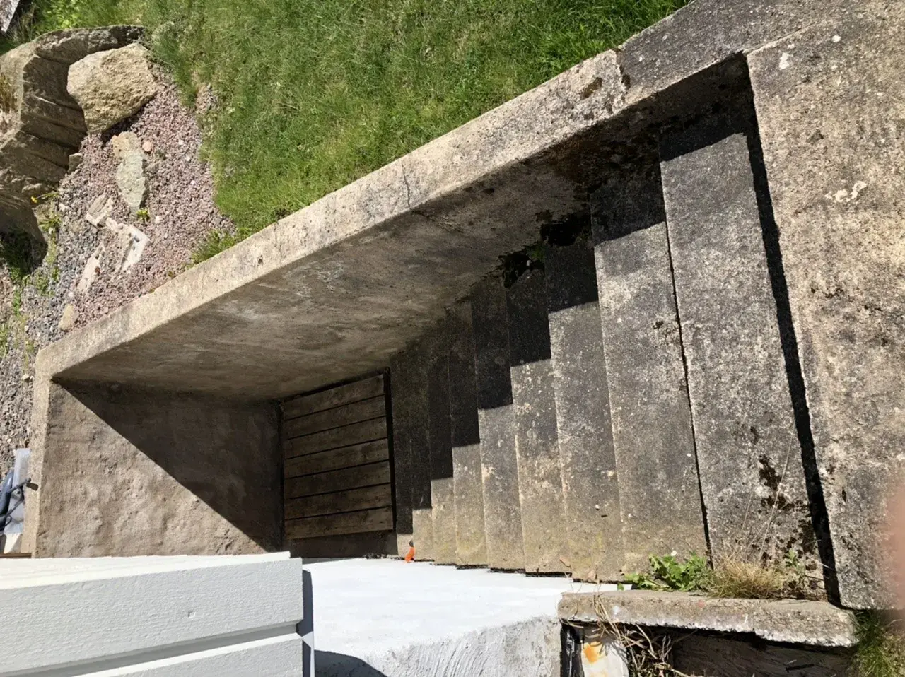 Betongtrappa leder ner till en träport. Gräs och stenar syns ovanför. Kanske någon ska borra i putsad fasad här snart.
