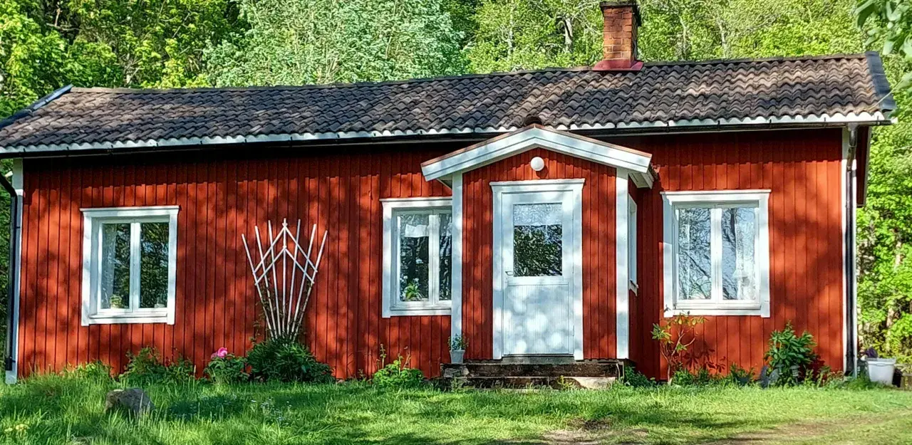 Röd stuga med vita knutar och tegeltak, omgiven av grönska. En klassisk bild som visar skillnad på torp och stuga.