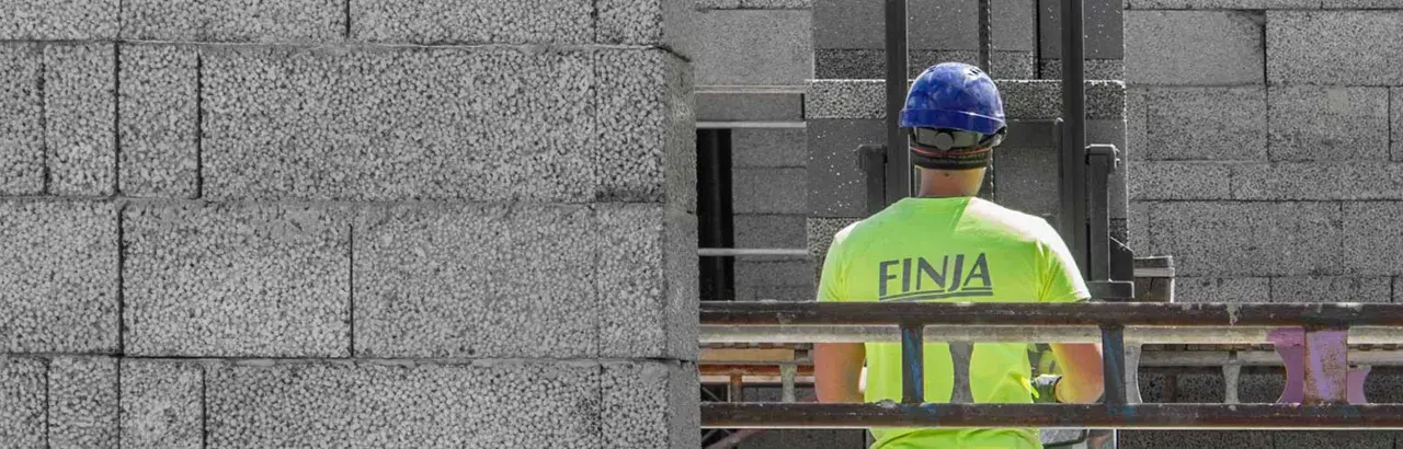 Construction worker in a bright green shirt with "FINJA" on the back, wearing a blue hard hat, next to a wall of concrete blocks.
