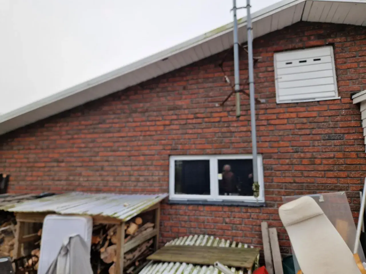Röd tegelfasad konstruktion med ett fönster och en vit garageport. Vedhögar och möbler syns framför huset.
