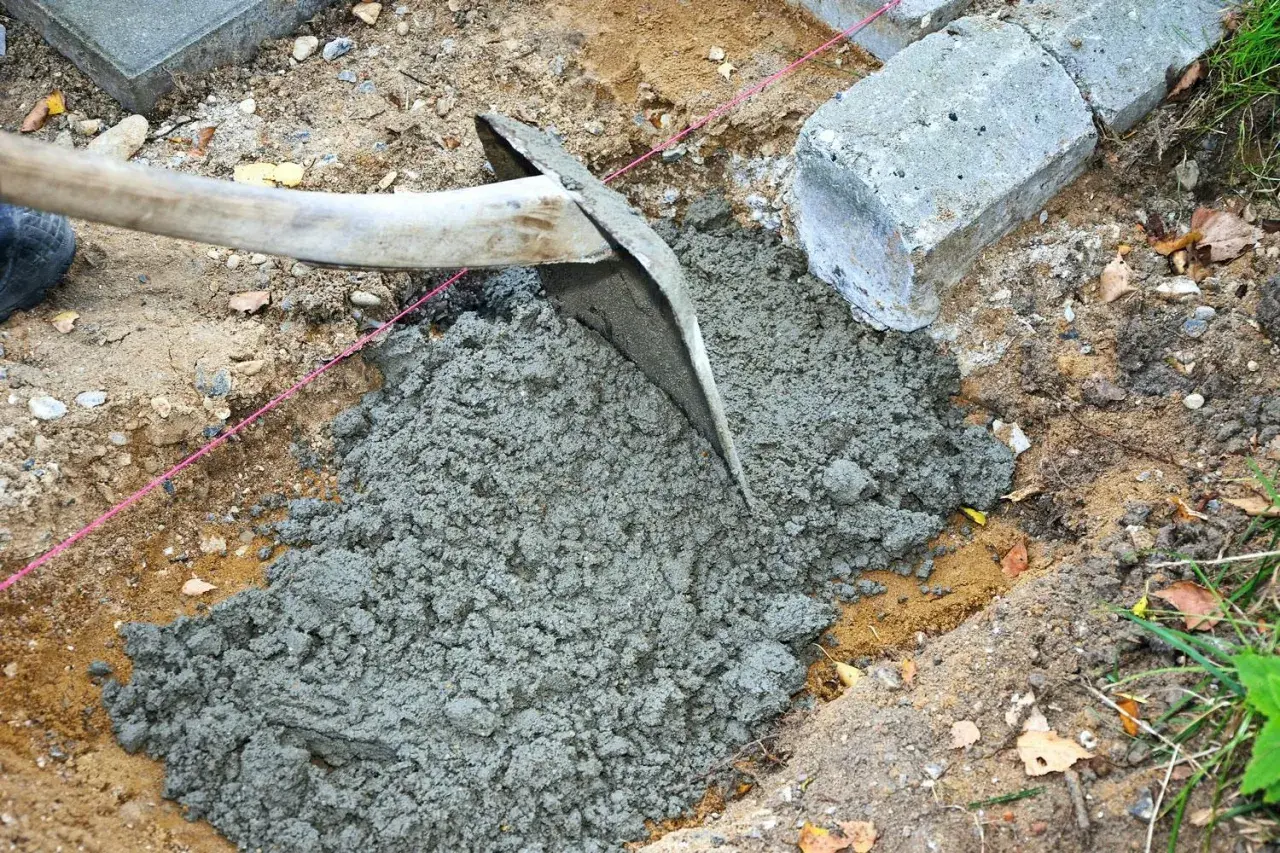 En hacka sprider jordfuktig betong i en grop. Betongblock och en rosa snodd syns i bakgrunden.