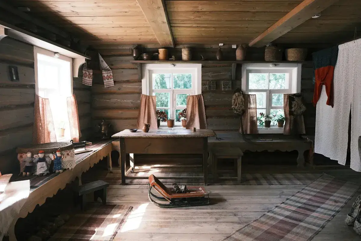 Interi&ouml;r i ett gammalt torp med tr&auml;v&auml;ggar, f&ouml;nster med blommor och dukar. En s&auml;ng med dockor och en sl&auml;de p&aring; golvet. En p&aring;minnelse om skillnad p&aring; torp och stuga.