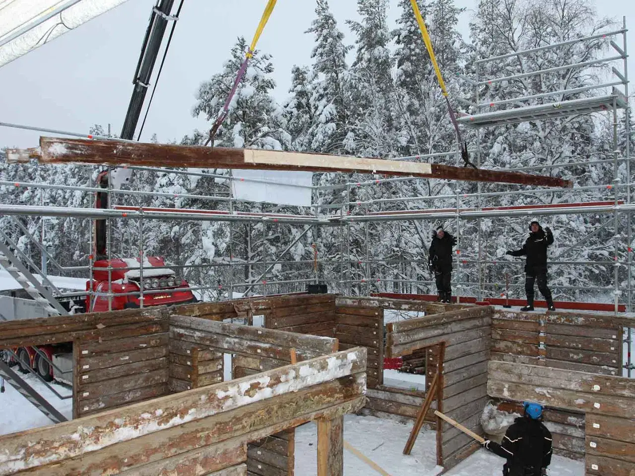 Arbetare flyttar timmerhusdelar med kran i snöig skogsmiljö. Bygget fortsätter trots kylan.