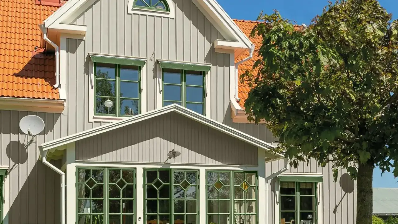 Ett charmigt ljusgr&aring;tt hus med orange takpannor och gr&ouml;na f&ouml;nsterkarmar. En stor l&ouml;nn skuggar delvis fasaden.