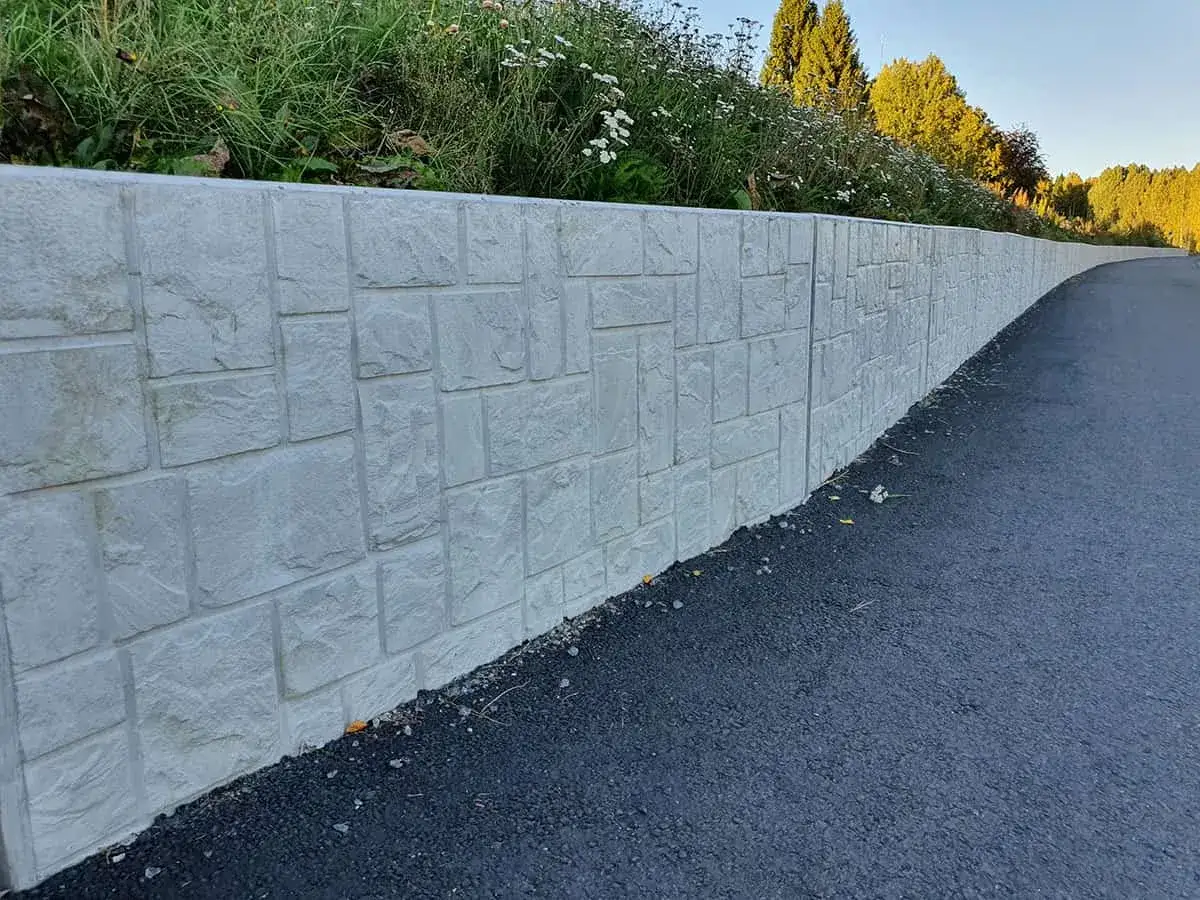 En nygjord betongmur med stenliknande m&ouml;nster l&auml;ngs en asfalterad v&auml;g. Gr&ouml;nskande v&auml;xter och tr&auml;d syns ovanf&ouml;r muren.