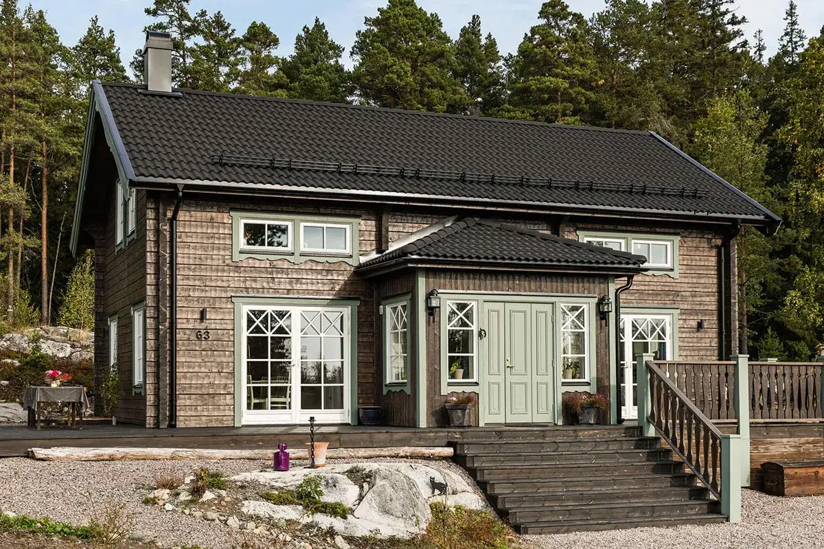 Vackert lgb timmerhus med gröna detaljer, omgivet av skog. En inbjudande entré med trappa leder upp till huset.