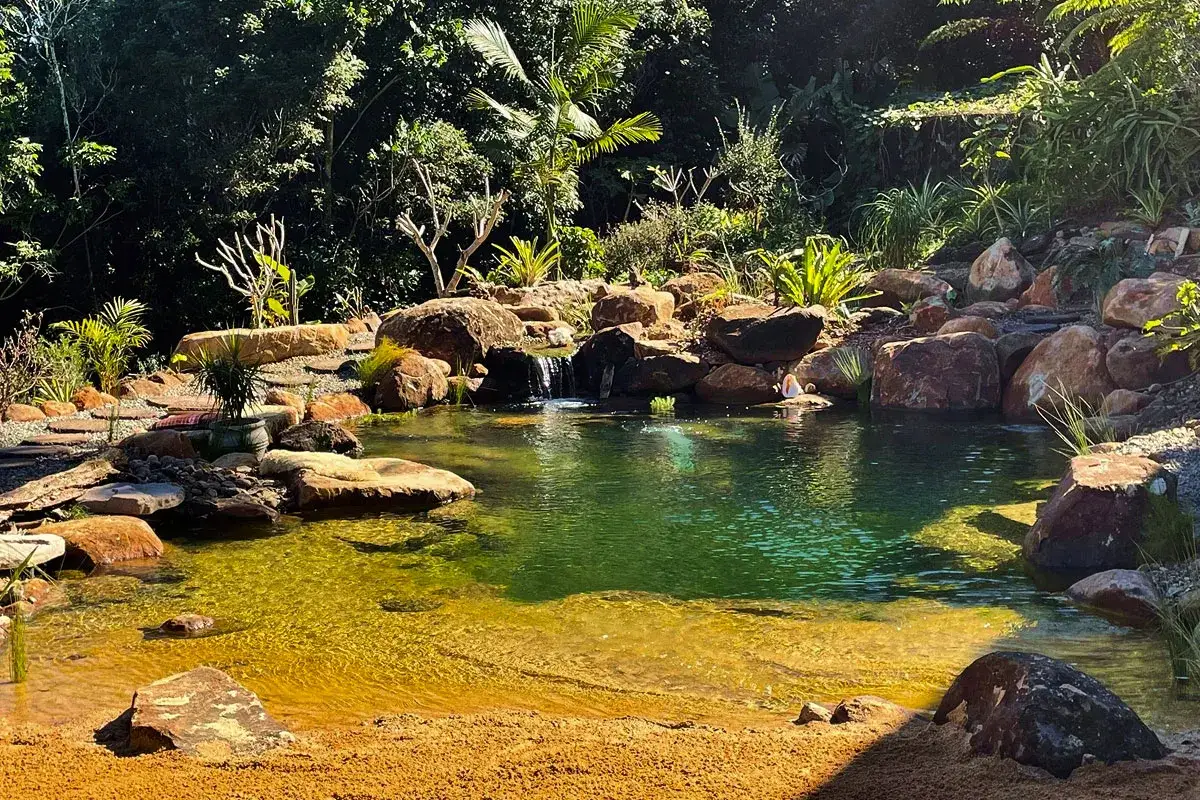 En vacker natur pool med ett litet vattenfall, omgiven av stenar och frodig gr&ouml;nska. En person sitter vid kanten.