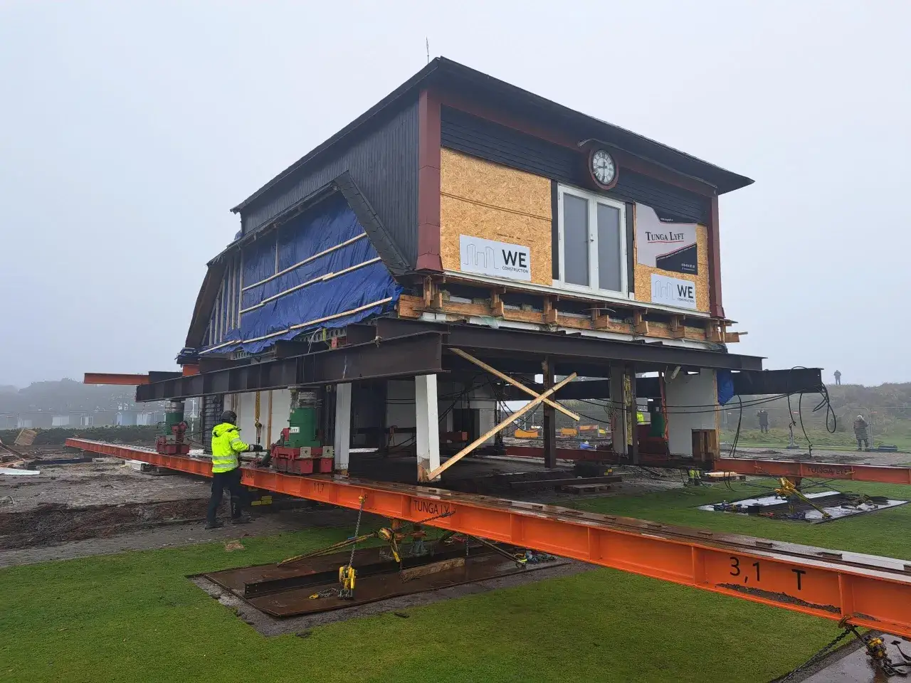 Ett hus lyfts med domkraft på orange balkar. En person i gul jacka övervakar arbetet.