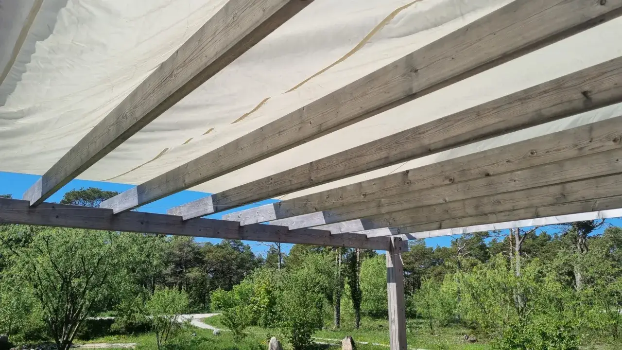 Träbjälkar stöder ett vitt pergolatak av tyg, som ger skugga över en grönskande trädgård med blå himmel.