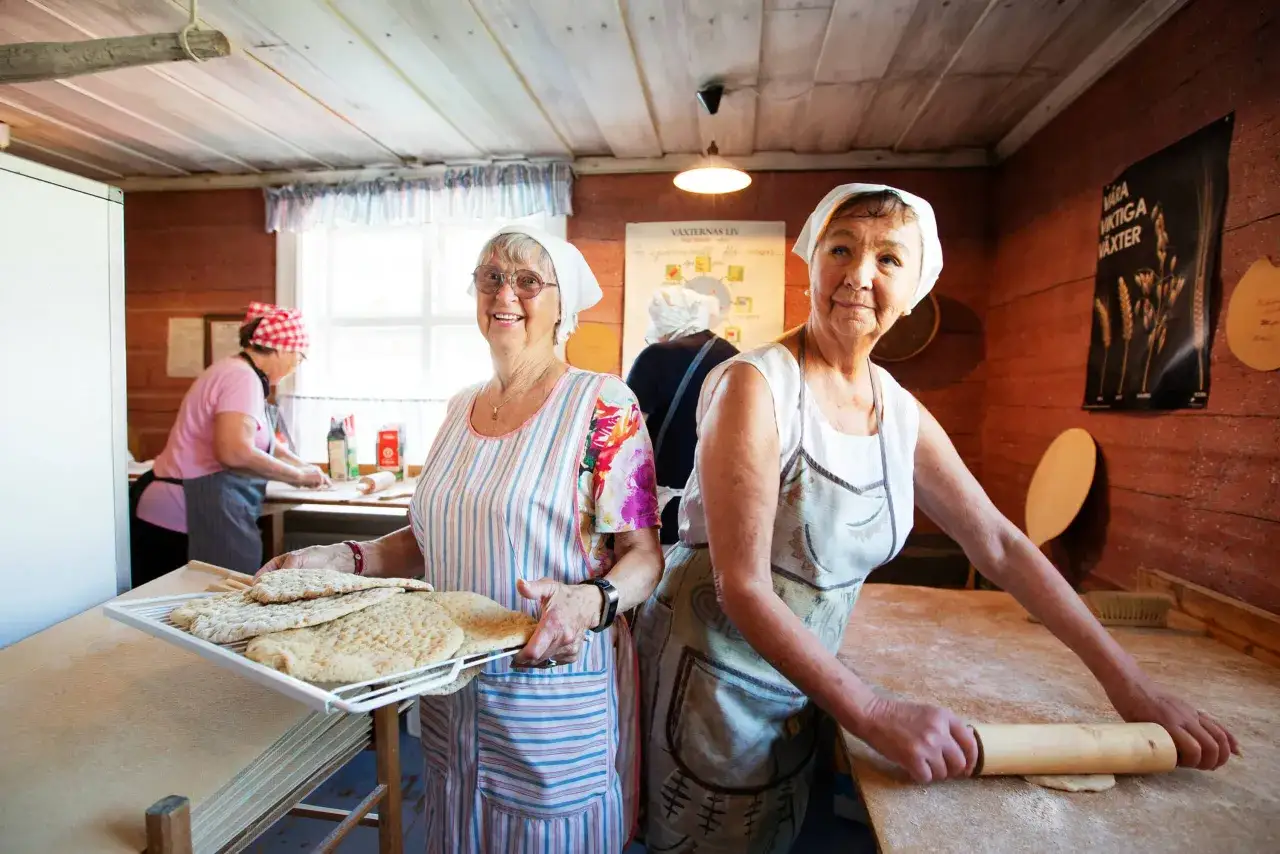 Kvinnor bakar tunnbr&ouml;d i en bagarstuga. En kvinna rullar deg, en annan h&aring;ller en bricka med nybakat br&ouml;d.
