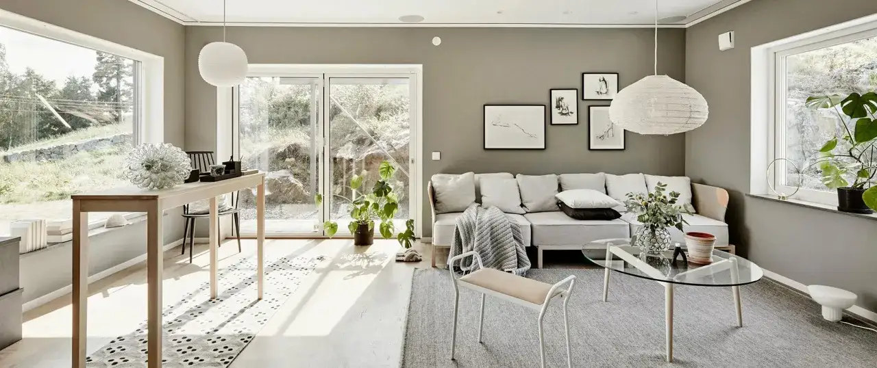Ljust vardagsrum med stora fönster som släpper in mycket ljus. En soffa, soffbord och ett långt träbord. En grön växt bidrar till känslan av hållbara hus.